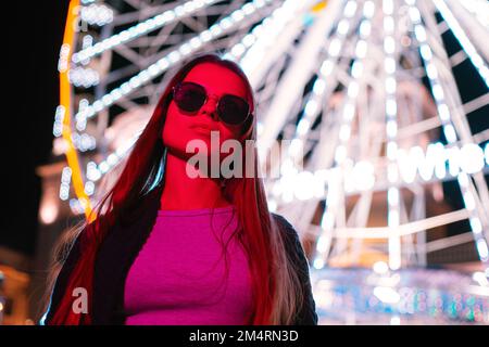 Ritratto una giovane donna urbana piuttosto turistica in occhiali da sole in piedi di fronte ad una ruota panoramica su sfondo rosso al neon di colore chiaro Foto Stock