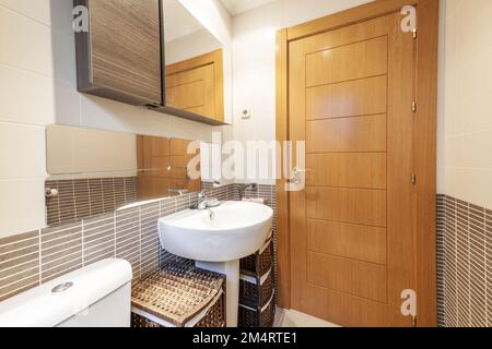 Bagno con mobili in rete di rattan, armadietto in legno con porta a specchio, lavabo in porcellana dal design profondo e porta in legno di pera Foto Stock