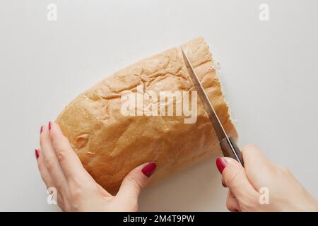 la mano femminile taglia un pezzo di pane con un coltello su un tavolo bianco in cucina a casa Foto Stock