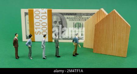 Concetto economico. Sulla superficie verde sono case di legno, una pila di dollari e figurine in miniatura di persone in piedi in linea per acquistare una casa. Foto Stock