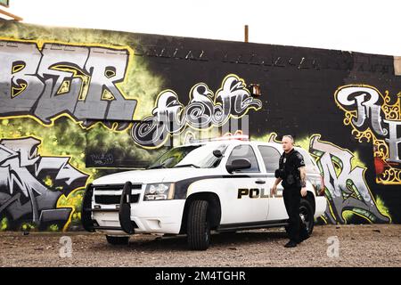 Immagine orizzontale di un poliziotto caucasico maschio bianco che esce dalla sua auto poliziotto nella sua città con graffiti sul muro come le auto backd Foto Stock