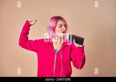 Una giovane bella ragazza in una blusa rosa con capelli luminosi ascoltando musica con gli auricolari collegati al telefono cellulare, chattando con gli amici mentre ballano, godendo la vita, su uno sfondo chiaro. Foto Stock