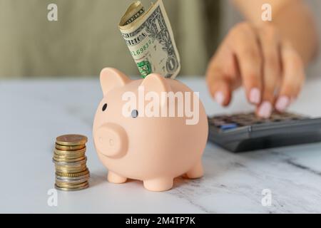 Primo piano della mano di una donna con una banca piggy e una calcolatrice sul tavolo dell'ufficio. Carta dollari e monete risparmio, crisi e inflazione. Gestione dei costi Foto Stock