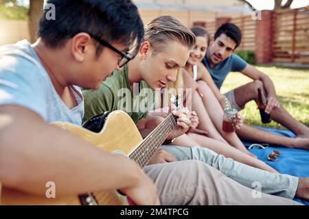 Strimpellimento via durante l'estate. un giovane che suona la chitarra mentre sorsegga qualche drink con i suoi amici fuori nel sole estivo. Foto Stock