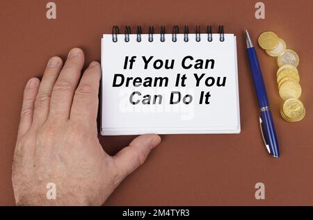 Concetto aziendale. Su una superficie marrone sono le monete, una penna e un blocco note con l'iscrizione - se si può sognare si può farlo. Il notebook sta tenendo Foto Stock