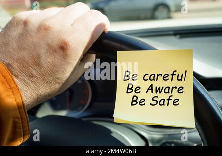 Il concetto di sicurezza dei trasporti. Un uomo tiene il volante accanto a un adesivo con la scritta Stai attento. Essere consapevoli. Essere al sicuro. Foto Stock