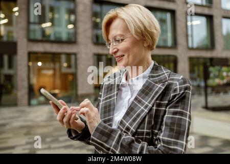 Donna d'affari matura sorridente negli occhiali usano il telefono mentre si trova in piedi sullo sfondo della costruzione della città Foto Stock