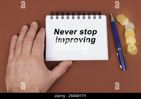 Concetto aziendale. Su una superficie marrone ci sono monete, una penna e un blocco note con l'iscrizione - non smettere mai di migliorare. Il notebook sta tenendo una mano. Foto Stock