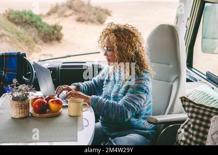 Una moderna turista che lavora su un computer portatile seduto all'interno di un moderno camper van con vista sulla spiaggia parcheggio in background. Donna che usa il computer in vehi Foto Stock