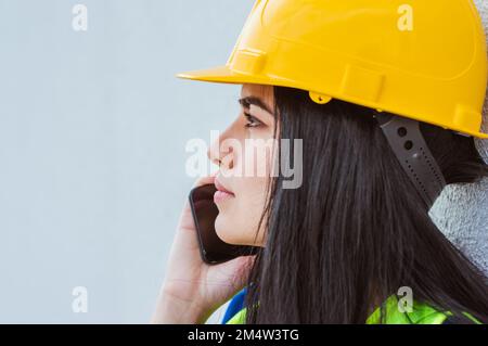 giovane donna venezuelana dai capelli lunghi ingegnere caucasico latina, indossando casco di sicurezza giallo e giubbotto, in piedi all'aperto appoggiato contro una parete, su un ph Foto Stock