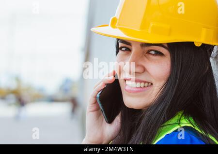 primo piano di giovane latina caucasica venezuelana ingegnere donna con casco giallo e giubbotto di sicurezza sta parlando al telefono, guardando la fotocamera sorridendo, Foto Stock