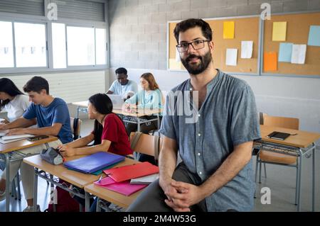 Ritratto di un insegnante maschio che guarda la macchina fotografica in una classe con gli studenti Foto Stock