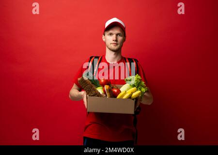 Il corriere in uniforme rossa contiene una scatola di carta per alimenti e generi alimentari. Servizio di consegna, ristoranti da asporto consegna cibo a casa. Foto Stock