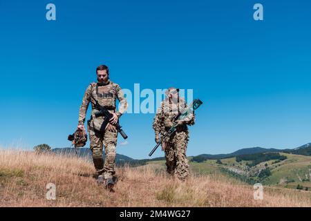 Una squadra di cecchini composta da soldati sta per essere coperta. Assistente Sniper e team leader camminando e puntando nella natura con erba gialla e cielo blu Foto Stock