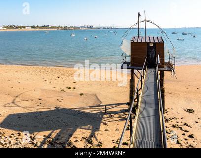 Una cabina di pesca in legno con una grande rete di ascensore quadrata chiamata 'carrelet' getta la sua ombra sulla spiaggia in bassa marea, di fronte al ponte di Saint-Nazaire. Foto Stock