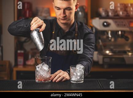 Il barman tiene in mano una paletta di metallo con cubetti di ghiaccio e li versa in vetro vecchio stile Foto Stock