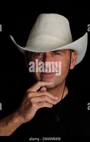 Un colpo verticale di un uomo sorridente con un dito sul mento indossando un cappello da fattoria su sfondo nero Foto Stock