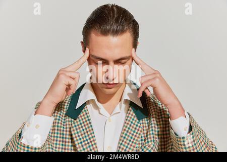 Giovane uomo triste e bello con capelli retrò in giacca vintage tenendo le dita sulla testa su sfondo grigio. Concetto di salute, emozioni Foto Stock