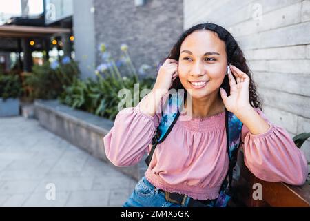 Donna studentessa dell'università latina sorridente e che usa gli auricolari seduti fuori dal centro commerciale nel tempo libero Foto Stock