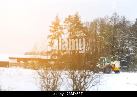 Defocus trattore pulito neve soleggiato bagliare. Il trattore pulisce la neve sul campo. Il grande trattore giallo pulisce la neve dalla strada e la carica nel camion. O Foto Stock