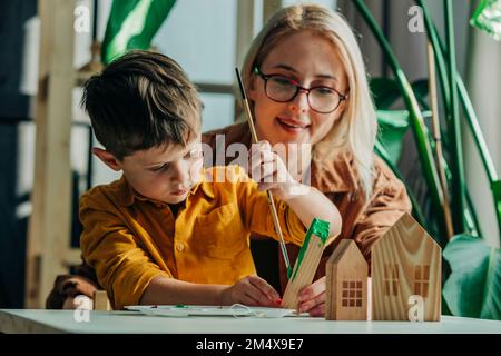 Sorridente madre e figlio dipingendo case modello in legno a tavola Foto Stock