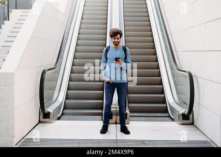 Giovane uomo che usa uno smartphone in piedi davanti alla scala mobile Foto Stock