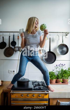 Donna matura sorridente in piedi sopra la stufa che si diverte in cucina Foto Stock