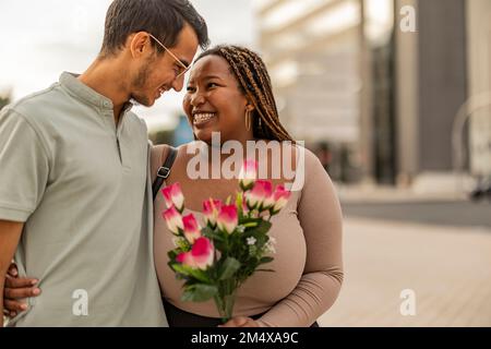 Felice giovane uomo abbracciando donna in piedi con bouquet di fiori Foto Stock