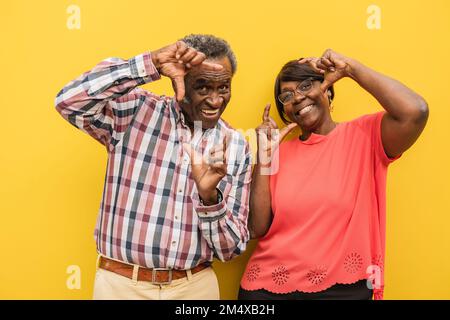 Coppia anziana felice che fa le cornici del dito contro lo sfondo giallo Foto Stock
