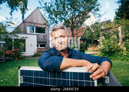 Uomo maturo sorridente seduto con pannello solare di fronte a casa Foto Stock