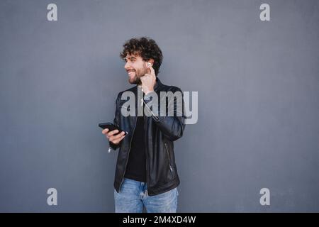 Uomo felice che tiene il telefono cellulare regolare le cuffie intrauricolari wireless davanti alla parete grigia Foto Stock