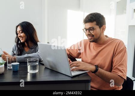 Uomo sorridente che studia attraverso un notebook seduto da una donna al tavolo nel soggiorno Foto Stock