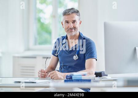 Medico sorridente seduto alla scrivania in pratica medica Foto Stock