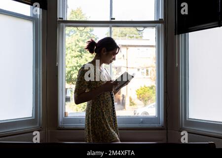 Donna sorridente che legge il libro in piedi davanti alla finestra Foto Stock
