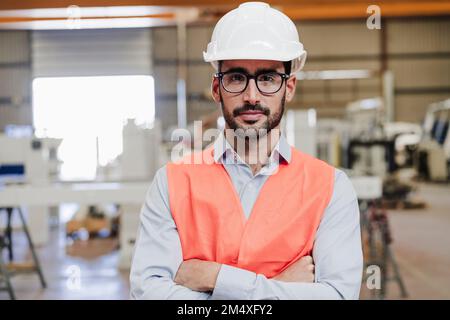 Ingegnere sicuro indossando occhiali e indumenti da lavoro protettivi in piedi con braccia incrociate nell'industria Foto Stock