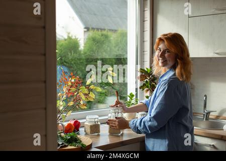 Donna sorridente in piedi in cucina alla finestra Foto Stock
