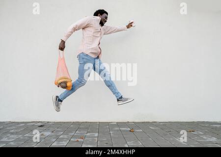 Uomo con borsa della spesa e tazza da caffè usa e getta che salta davanti alla parete bianca Foto Stock