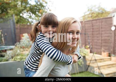 Ragazza sorridente che dà cavalcata piggyback a sorella in giardino Foto Stock