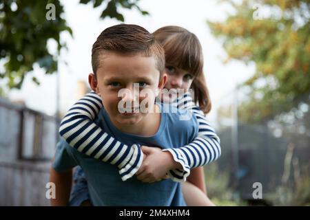 Ragazzo giocoso che dà cavalcata piggyback a sorella in giardino Foto Stock
