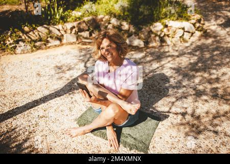 Donna matura felice che pratica yoga con smartphone nel cortile posteriore Foto Stock