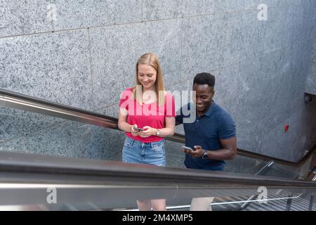 Sorridendo uomo e donna usando smartphone che si spostano su scala mobile a parete Foto Stock