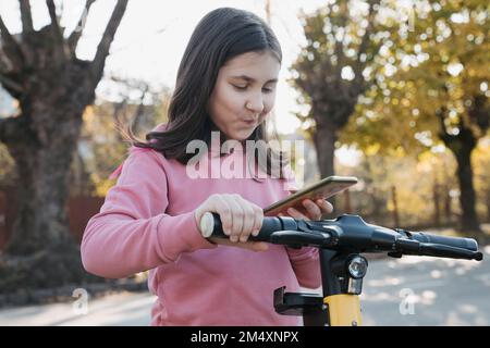 Ragazza che usa lo smartphone per noleggiare uno scooter elettrico Foto Stock