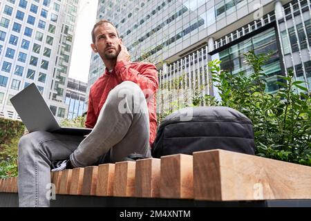 Uomo seduto con un computer portatile davanti agli edifici moderni Foto Stock