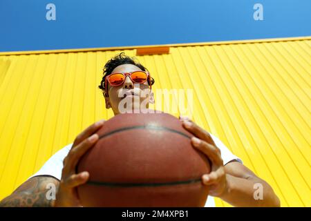 Uomo che indossa occhiali da sole rossi che tengono il basket davanti alla parete gialla Foto Stock