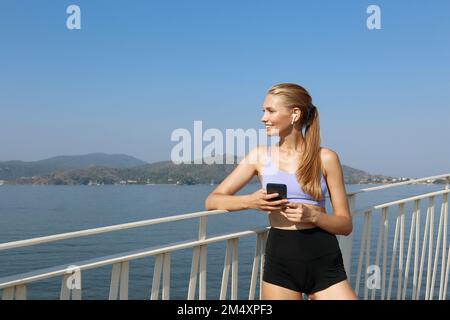 Donna sorridente che ascolta la musica con cuffie auricolari wireless che tengono il telefono cellulare appoggiato sulla ringhiera Foto Stock