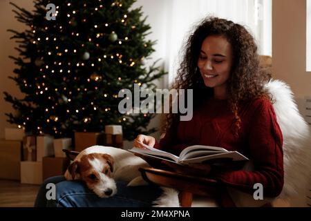 Donna felice seduta con il cane e libro di lettura a casa Foto Stock