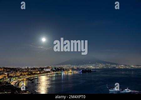 Vista sul golfo di Napoli in Italia, il lungomare in primo piano e il Vesuvio sullo sfondo. La luna piena sorge sopra l'orizzonte. Foto Stock