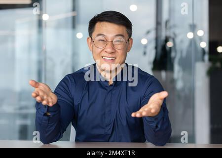 Un bel giovane asiatico è seduto in ufficio, guardando la macchina fotografica, puntando la macchina fotografica, sorridendo. Blogger. riunione online. formazione aziendale, insegnante. Foto Stock