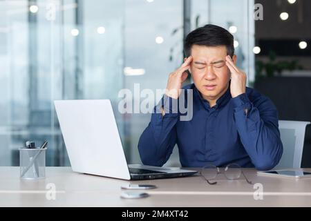 Stanco giovane uomo d'affari asiatico sente grave mal di testa emicrania. Si siede alla sua scrivania tiene la testa con le mani chiude gli occhi Foto Stock