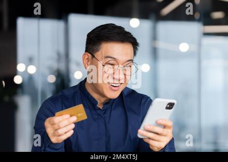 Primo piano. Un giovane uomo asiatico felice in occhiali tiene una carta di credito e guarda con gioia il telefono. Controlla l'account, effettua acquisti online, ordini. Foto Stock